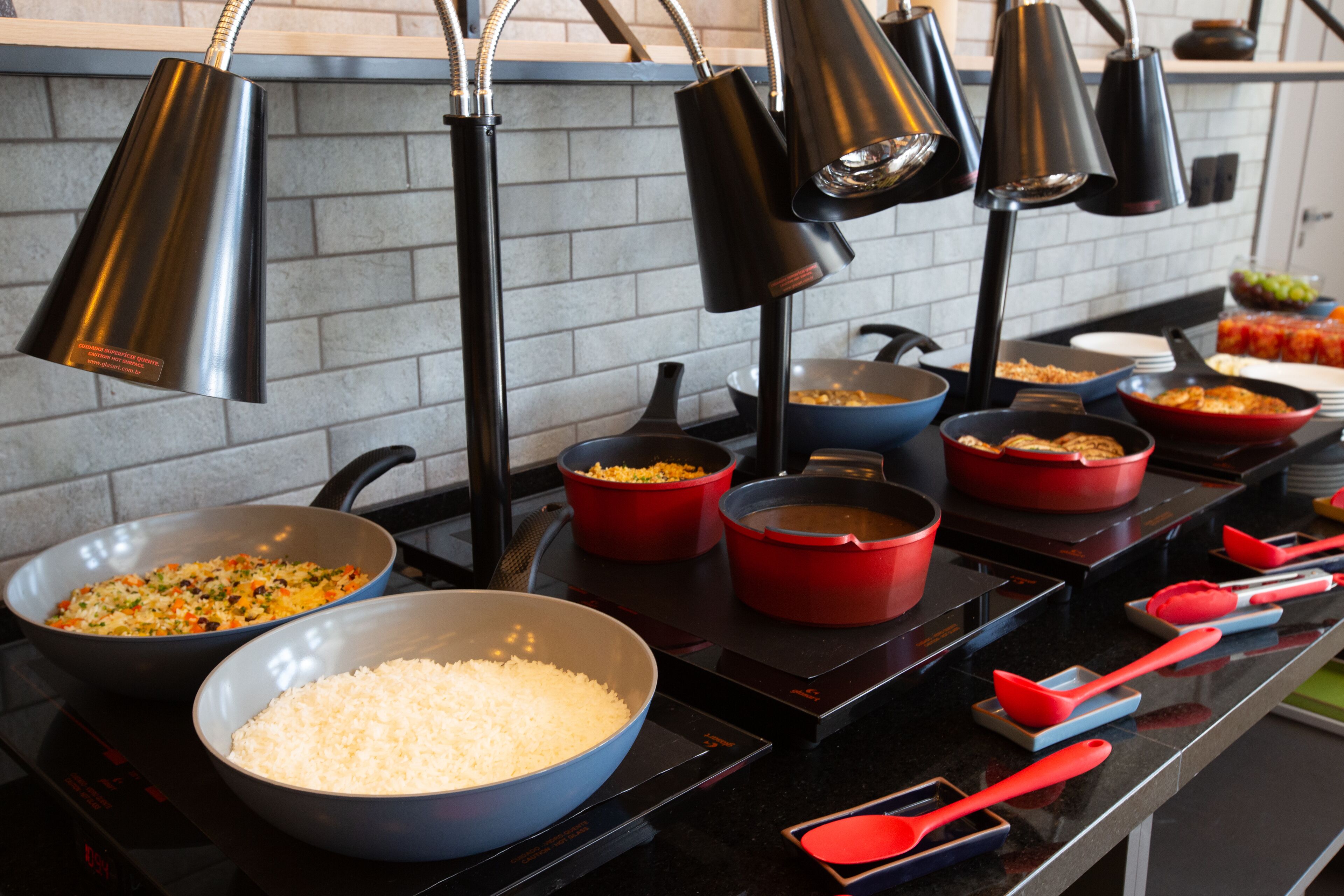 Uma bancada de cozinha com várias panelas e frigideiras, um relógio digital e utensílios de cozinha vermelhos.