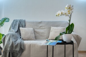 Una habitación con un sofá color beige acolchado, una manta gris, una orquídea blanca en maceta blanca y una revista sobre una mesita auxiliar negra.