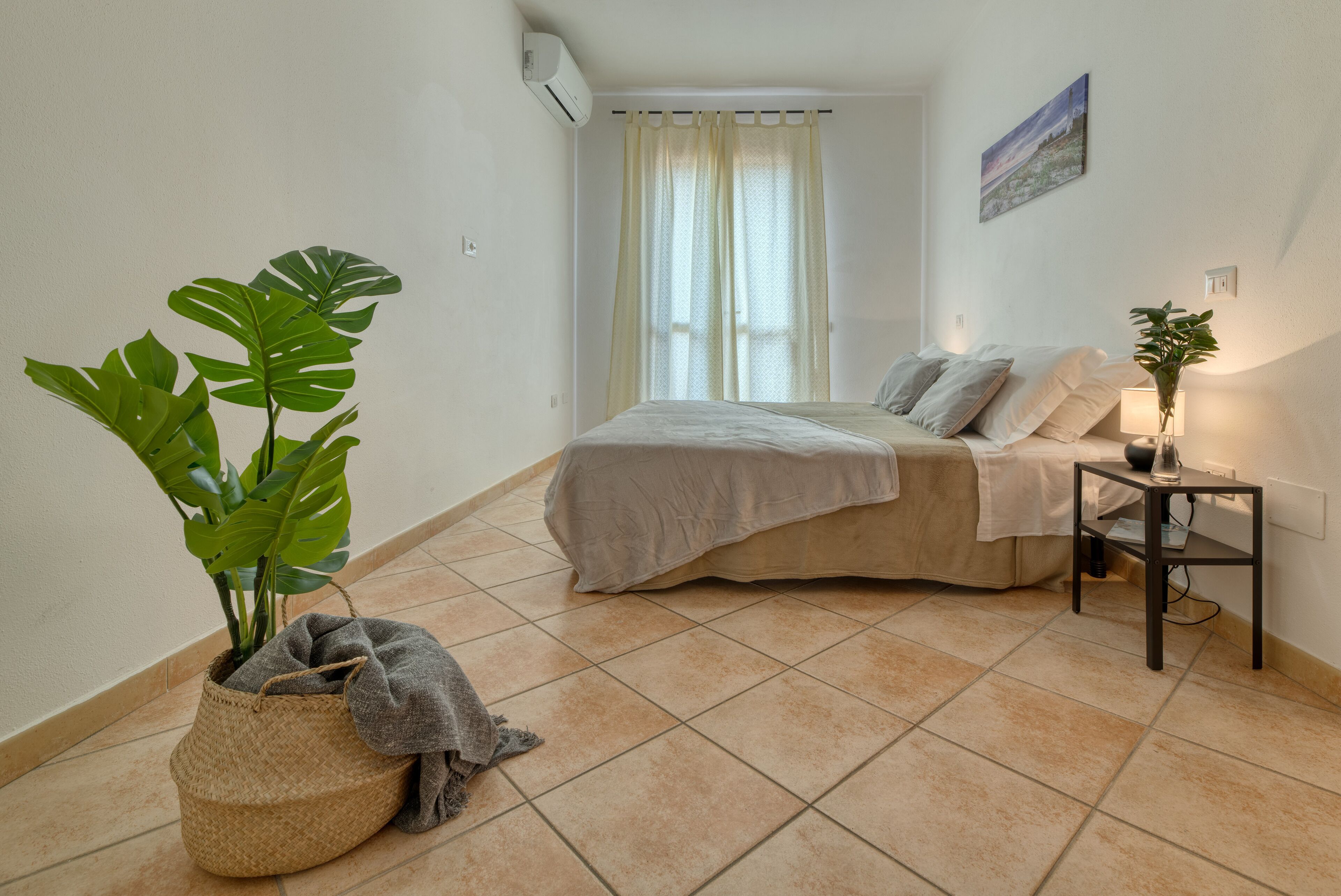 Un dormitorio con cama, una mesita de noche con lámpara, una planta en maceta y una ventana con cortinas.