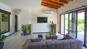 A modern living room with a large sofa, a glass coffee table, and a television mounted on the wall. There are two potted plants and a wooden column with a stack of logs.