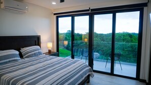A bedroom with a large bed, a nightstand, a lamp, and a view of a balcony with chairs and a table.