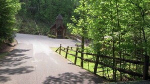 A rural road leading to a small wooden building with a sloped roof, surrounded by dense green foliage. - Countryside in Country Pines (Sevierville)