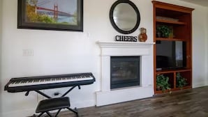 A living room with a fireplace, a piano, a framed picture of the Golden Gate Bridge, and a mirror with the word "CHEERS" above it.