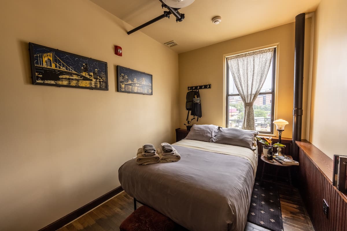 A hotel room with a large bed, two paintings on the wall, a window with a curtain, a bedside table with a lamp, and a coat rack.