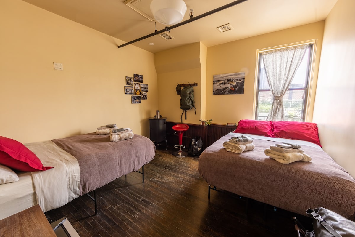 A room with two beds, a red stool, a coat rack, and a window with curtains.