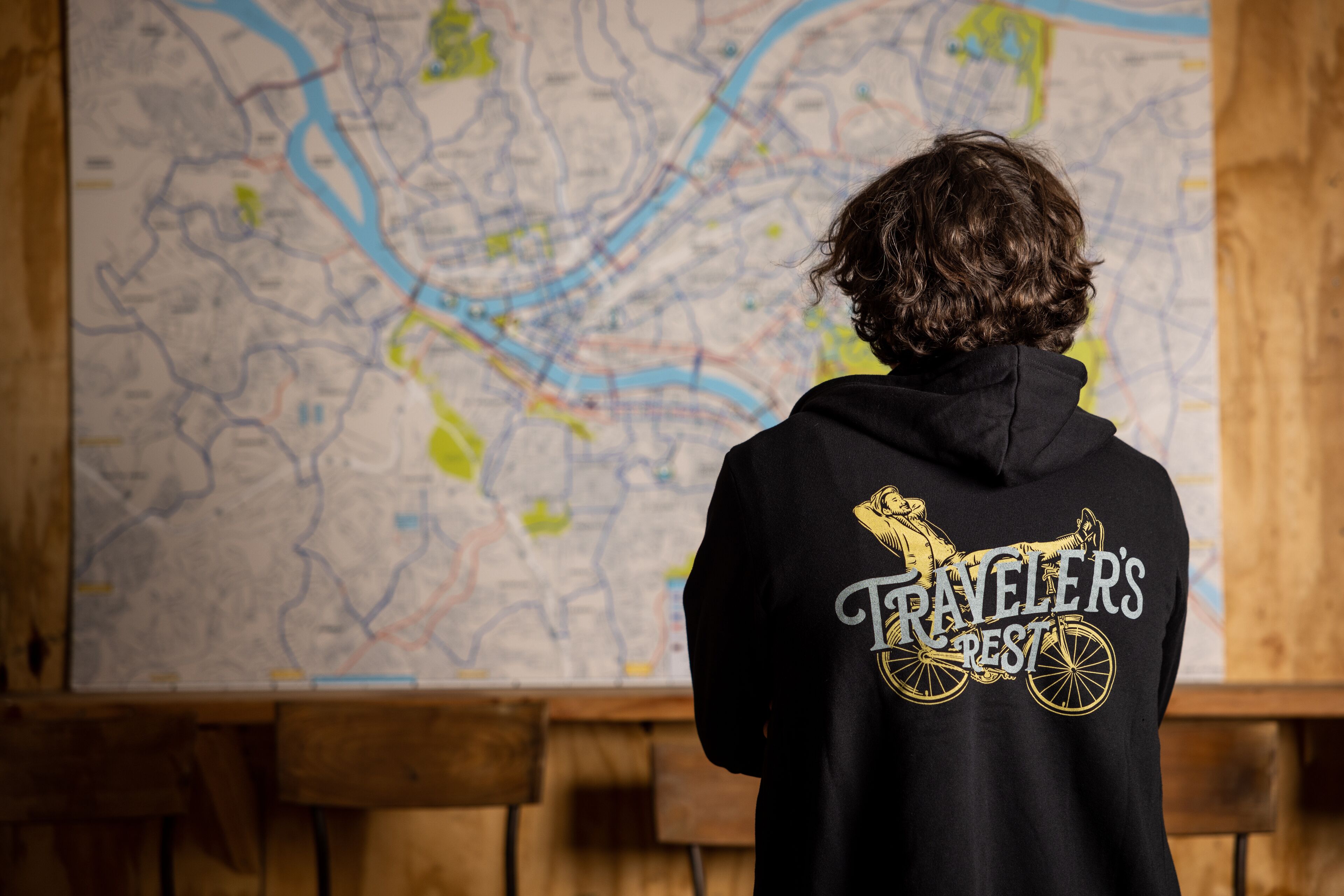 Una persona con una sudadera negra con un diseño gráfico y texto en la espalda, de pie frente a un mapa grande.