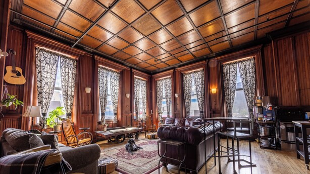 A spacious living room with a wooden ceiling, large windows with curtains, a leather sofa, a dining table, and a kitchen area. - Traveler's Rest Hotel (Pittsburgh)