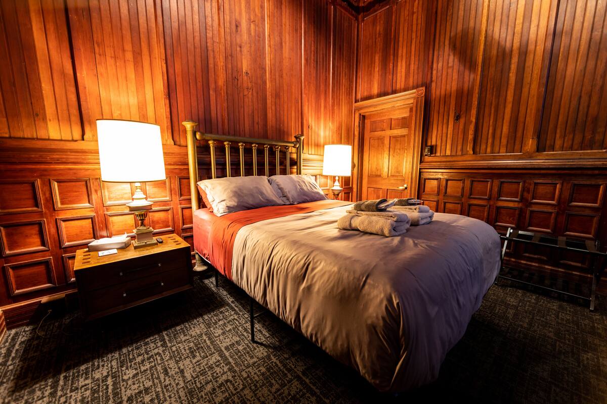 A wooden-paneled room with a bed, two bedside lamps, a nightstand, and a dresser.