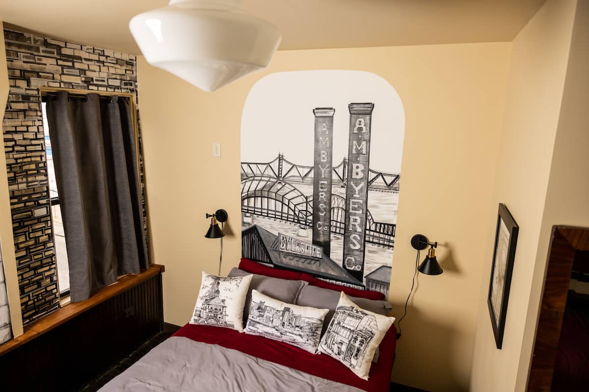 A bedroom with a bed, a wall-mounted lamp, a framed picture, and a window with curtains.