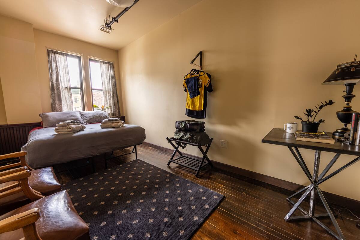 A hotel room with a bed, a desk, a chair, a rug, and a jersey hanging on the wall.