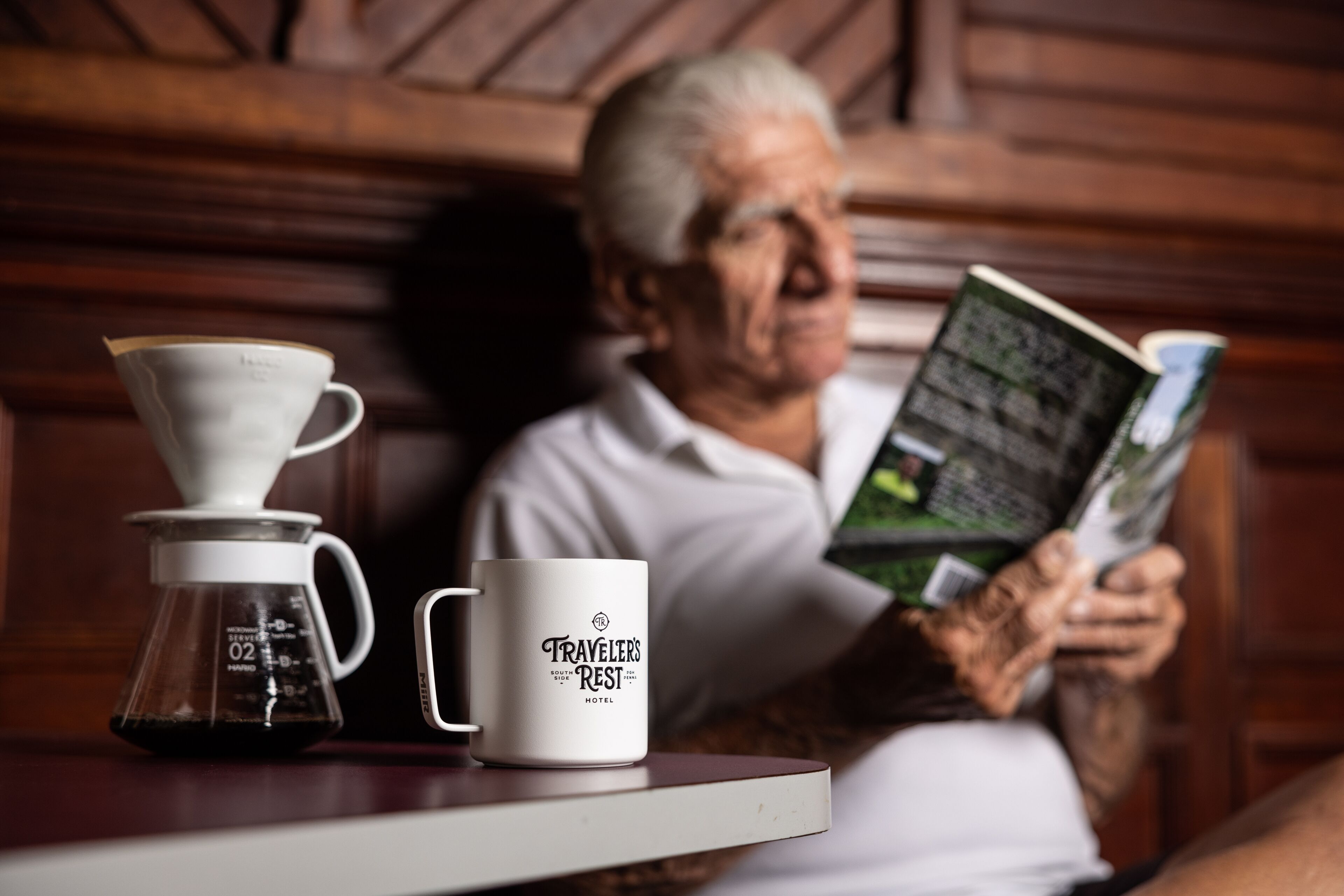 Une machine à café et une tasse sur une table, avec une personne qui lit un livre.