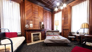 A room with a bed, a chair, a table, a lamp, and a fireplace. - Traveler's Rest Hotel (Pittsburgh)