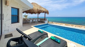 A poolside area with lounge chairs and a thatched umbrella, overlooking a clear blue sea.