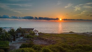 Een kustlandschap met een ondergaande zon, een strand en een huis met omheinde tuin.