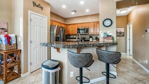 A kitchen with wooden cabinets, a stainless steel refrigerator, a microwave, and a granite countertop. There are two bar stools and a trash can. - NEW! Cozy desert getaway with private backyard and seasonal pool! (New River)