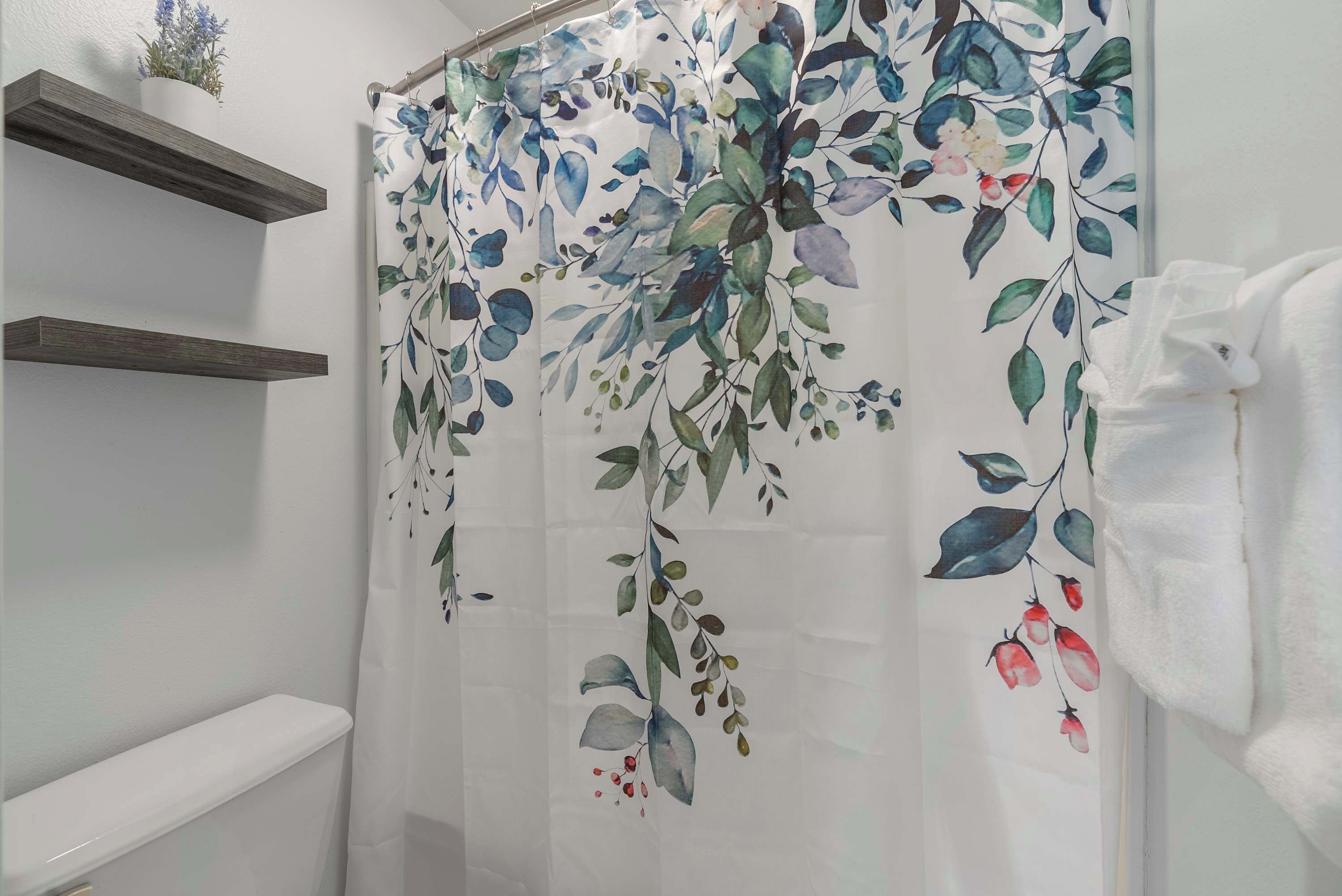 Un baño con cortina de ducha estampada con motivos florales, estantes de madera y una toalla blanca.