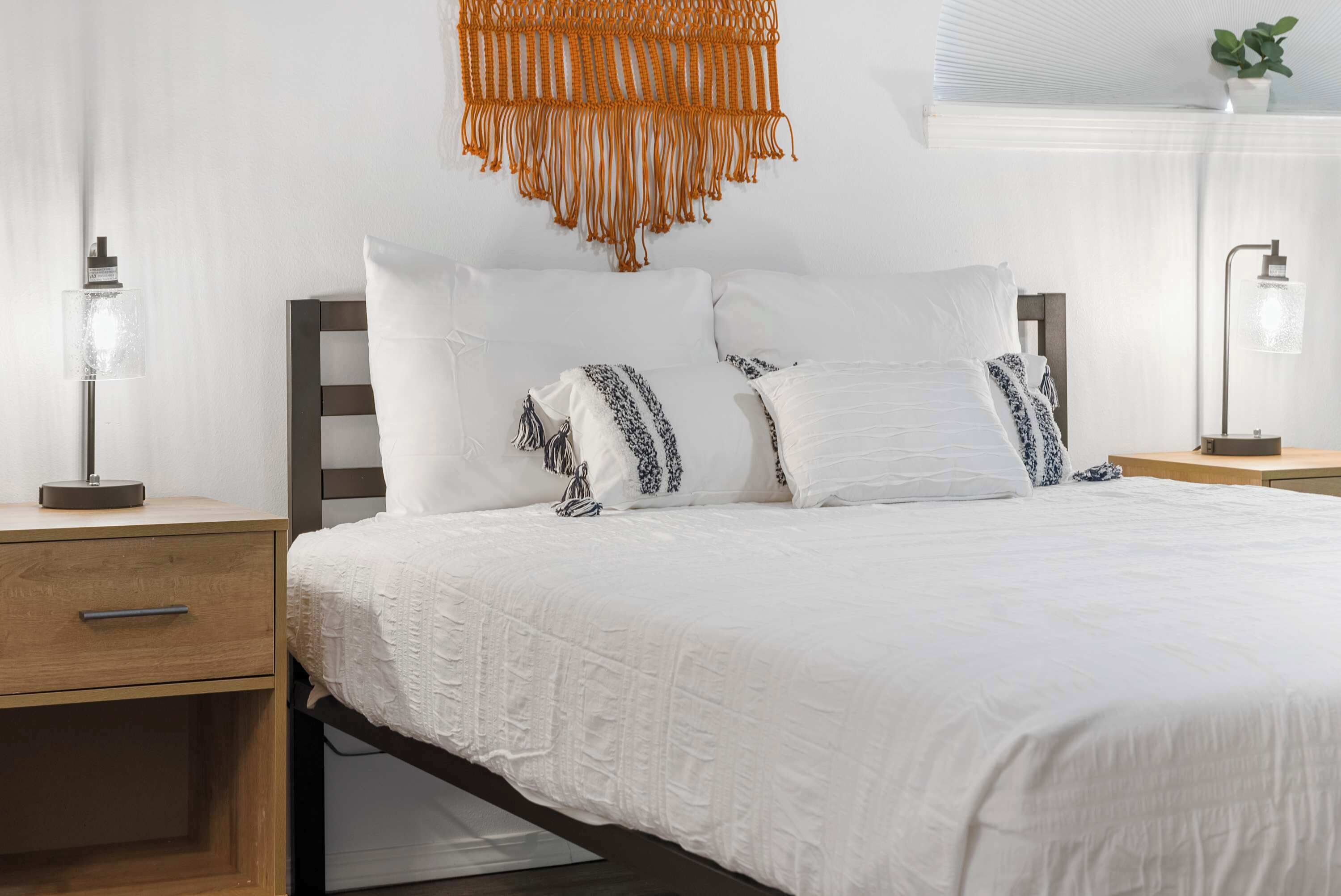 A neatly made bed with white bedding and patterned pillows, a wooden nightstand with a lamp, and a macrame wall hanging.