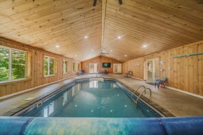 Piscina interna com paredes e teto de madeira, ventilador de teto e televisão fixada na parede.