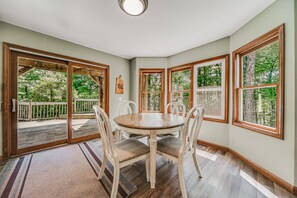 Uma área de refeições com mesa redonda e cadeiras, janelas amplas com vista para árvores e um deck de madeira na área externa.