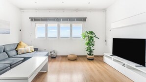 Un salón moderno con un sofá gris, una mesa de centro blanca, un televisor de pantalla plana grande, suelo de madera y un ventanal con vistas.