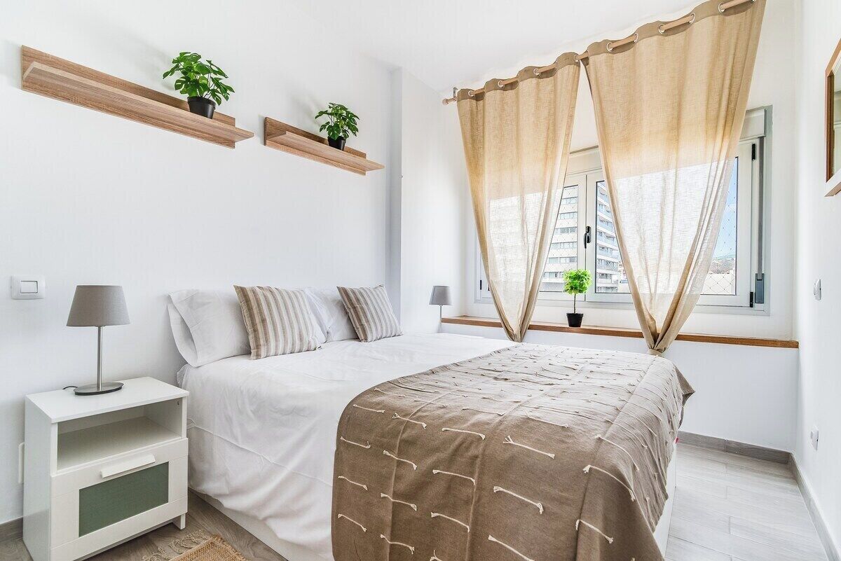 Un dormitorio con cama, mesitas de noche, una ventana con cortinas y estantes en la pared con plantas.