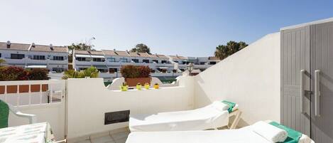 A rooftop terrace with two sun loungers, a table, and a view of a row of buildings.