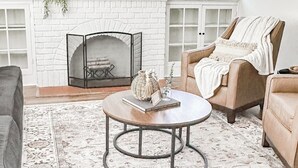 Una sala de estar con chimenea de ladrillo blanco, un televisor de pantalla plana montado arriba y un sillón beigue con una manta.