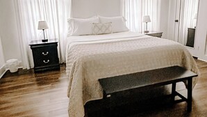 Un dormitorio con una cama, mesitas de noche, un banco y una ventana con cortinas.