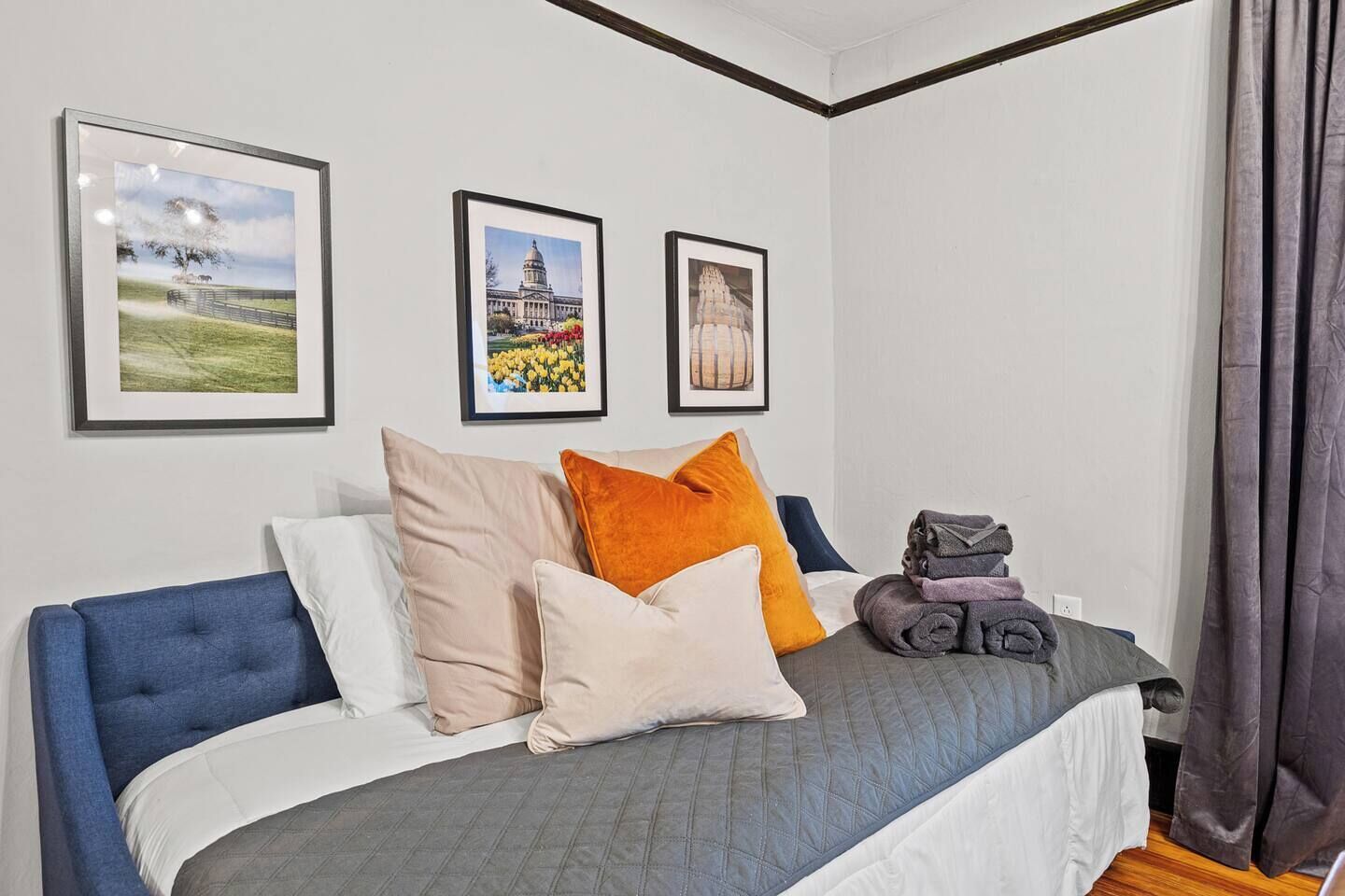 Un dormitorio con una cama, almohadas, una colcha y una pila de toallas dobladas. Hay tres cuadros en la pared.