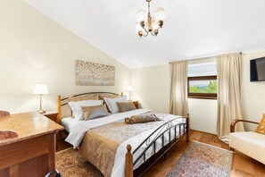 Een slaapkamer met een bed, nachtkastjes, een bureau, een stoel, een kroonluchter, een raam met gordijnen en een schilderij aan de muur.