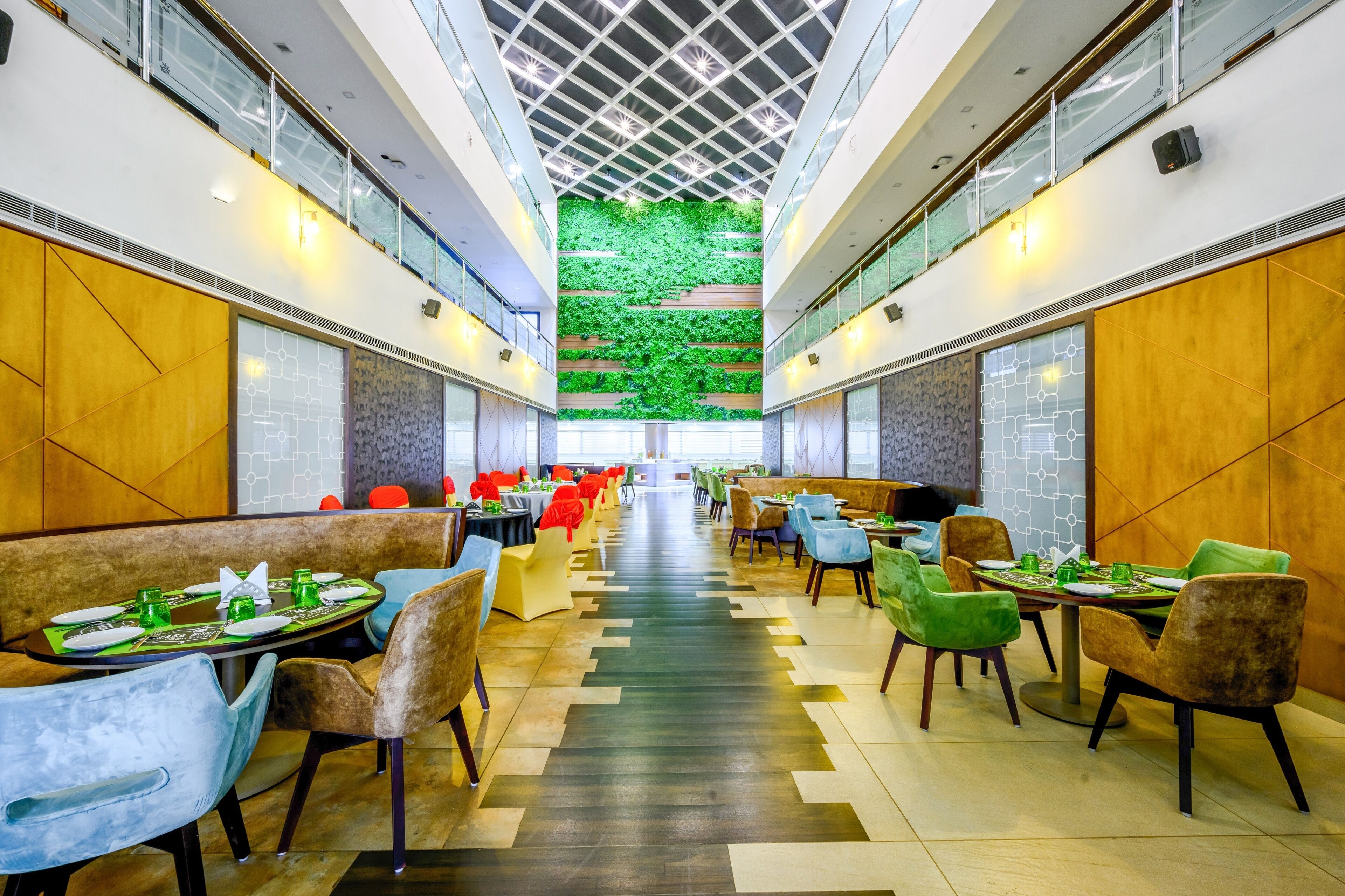 A spacious indoor dining area with multiple tables and chairs, a central green wall, and a high ceiling with a geometric pattern.