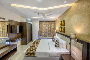 A modern hotel room with a large bed, a flat-screen TV mounted on the wall, a ceiling fan, and a window with patterned curtains. - Ranjitha Palace (Manipal Inn) (Udupi)