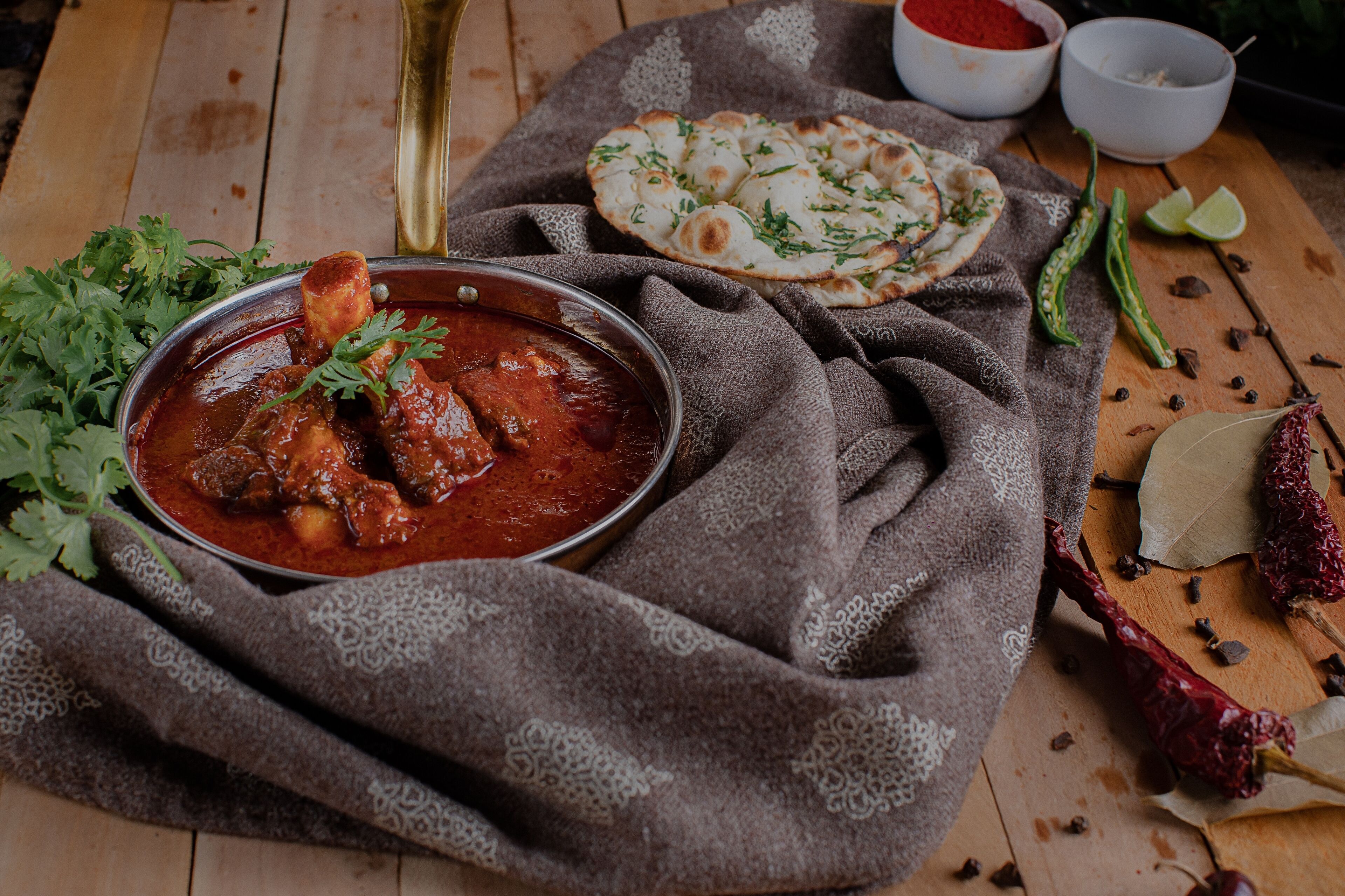 A meal with a bowl of stew, naan bread, and fresh herbs on a wooden table.