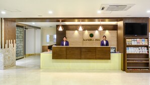 A reception desk with a wooden counter and two receptionists in suits. There is a wall-mounted TV and a bookshelf with magazines. - Ranjitha Palace (Manipal Inn) (Udupi)