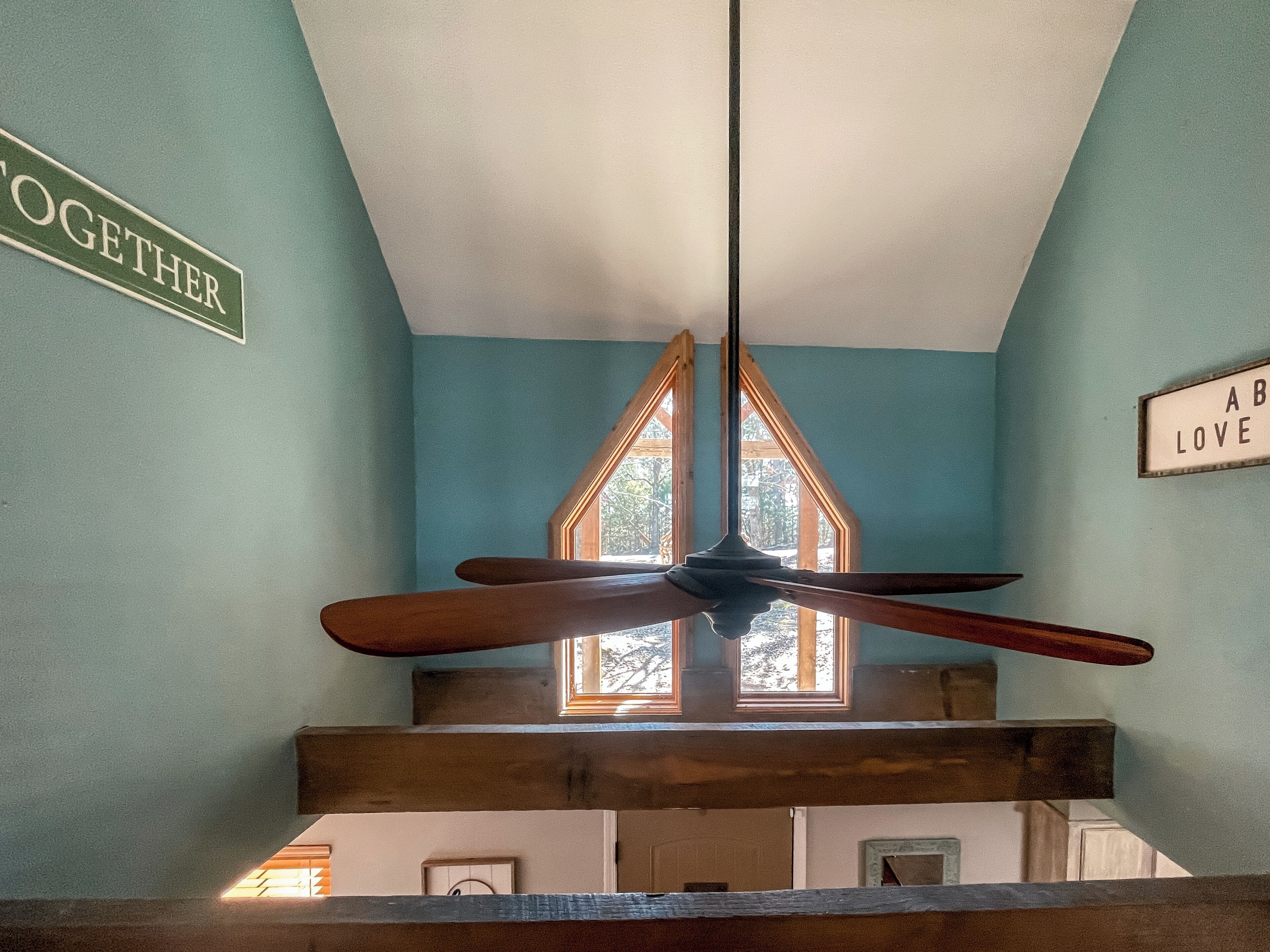 Un ventilador de techo en una habitación con paredes color turquesa, estructura de vigas de madera y una ventana con vista a árboles.