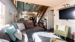 A cozy living room with a sofa, a green armchair, and a coffee table with a plant. A staircase leads to a kitchen area with a dining table and chairs. - Wister Ridge Cottage • Sleeps 10 • 3 Minutes to Wister Lake! (Wister)