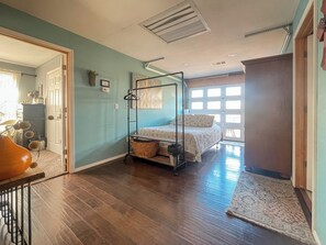 A room with a bunk bed, a wooden floor, a door leading to another room, and a window with blinds. - Wister Ridge Cottage • Sleeps 10 • 3 Minutes to Wister Lake! (Wister)