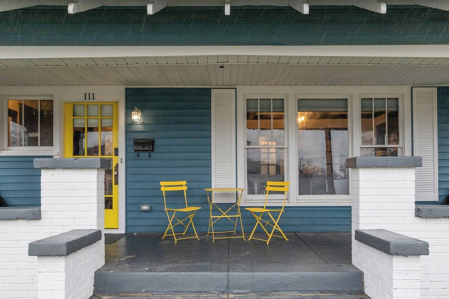 A house with a porch, numbered 111, featuring a yellow door, two windows with shutters, and a small table with yellow chairs.