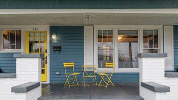 A house with a porch, numbered 111, featuring a yellow door, two windows with shutters, and a small table with yellow chairs.