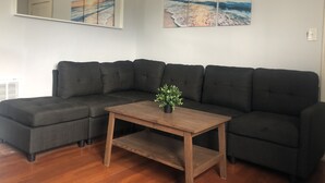 Un salon avec un canapé d’angle gris foncé, une table basse en bois ornée d’une plante et deux tableaux accrochés au mur représentant une scène de plage.