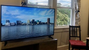Un téléviseur diffusant une image d’un paysage urbain avec un plan d’eau, placé devant une fenêtre ornée de rideaux.
