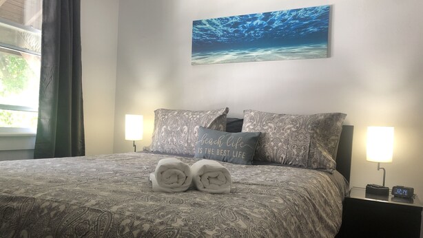 Un lit bien fait, recouvert d’une couvre-lit à motifs, avec deux oreillers et deux serviettes enroulées posées sur le lit. Au-dessus du lit, un tableau représentant une scène sous-marine est accroché au mur.