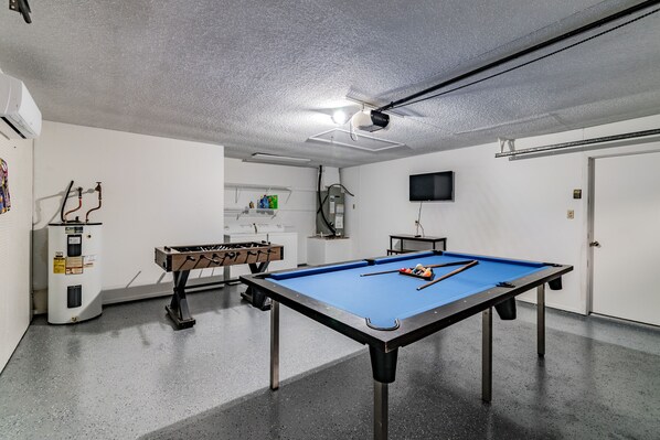 Una mesa de billar con superficie de fieltro azul, una mesa de fútbol de mesa y un televisor colgado en la pared.