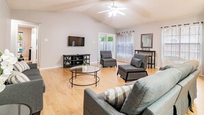 A spacious living room with a ceiling fan, a TV mounted on the wall, and a large window with blinds. - Entire House with Game Room in Lutz, Near Tampa and Wesley Chapel (Lutz)