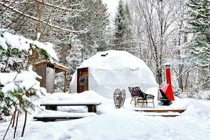 Ein verschneiter Außenbereich mit einer Holzhütte, einer verschneiten Bank und einem roten Schornstein.