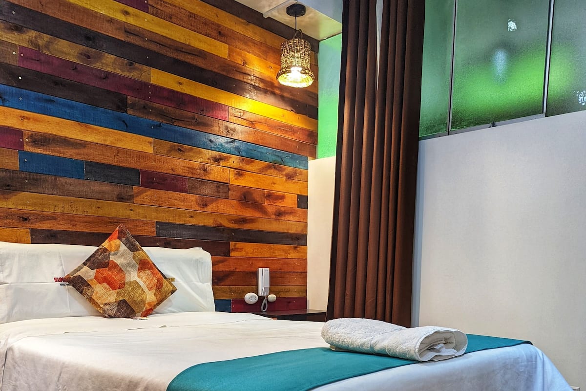 Cama bem arrumada com manta azul-esverdeada, almofada estampada e toalha branca dobrada ao lado. O quarto apresenta painéis de parede em madeira, telefone sobre uma mesa pequena e piso estampado.