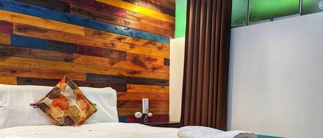 Una cama cuidadosamente tendida con un plaid verde azulado, una almohada estampada y una toalla blanca doblada al lado. La habitación presenta un diseño de paneles de madera, un teléfono sobre una mesita y un suelo estampado.