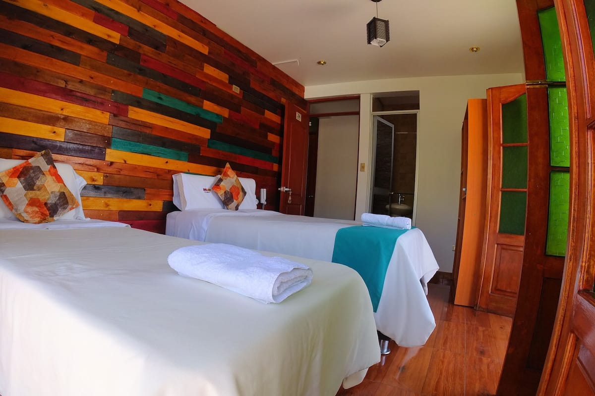 Quarto de hotel com duas camas, cabeceiras de ripas de madeira e um banheiro visível por uma porta aberta.