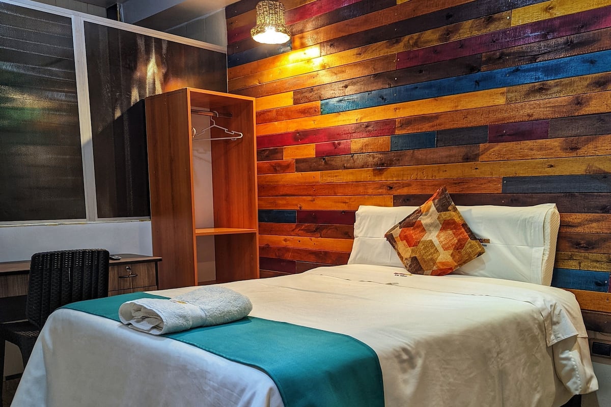 Cama bem arrumada com manta azul-turquesa, travesseiro estampado e edredom branco. Um guarda-roupa de madeira e uma cadeira estão visíveis no quarto.