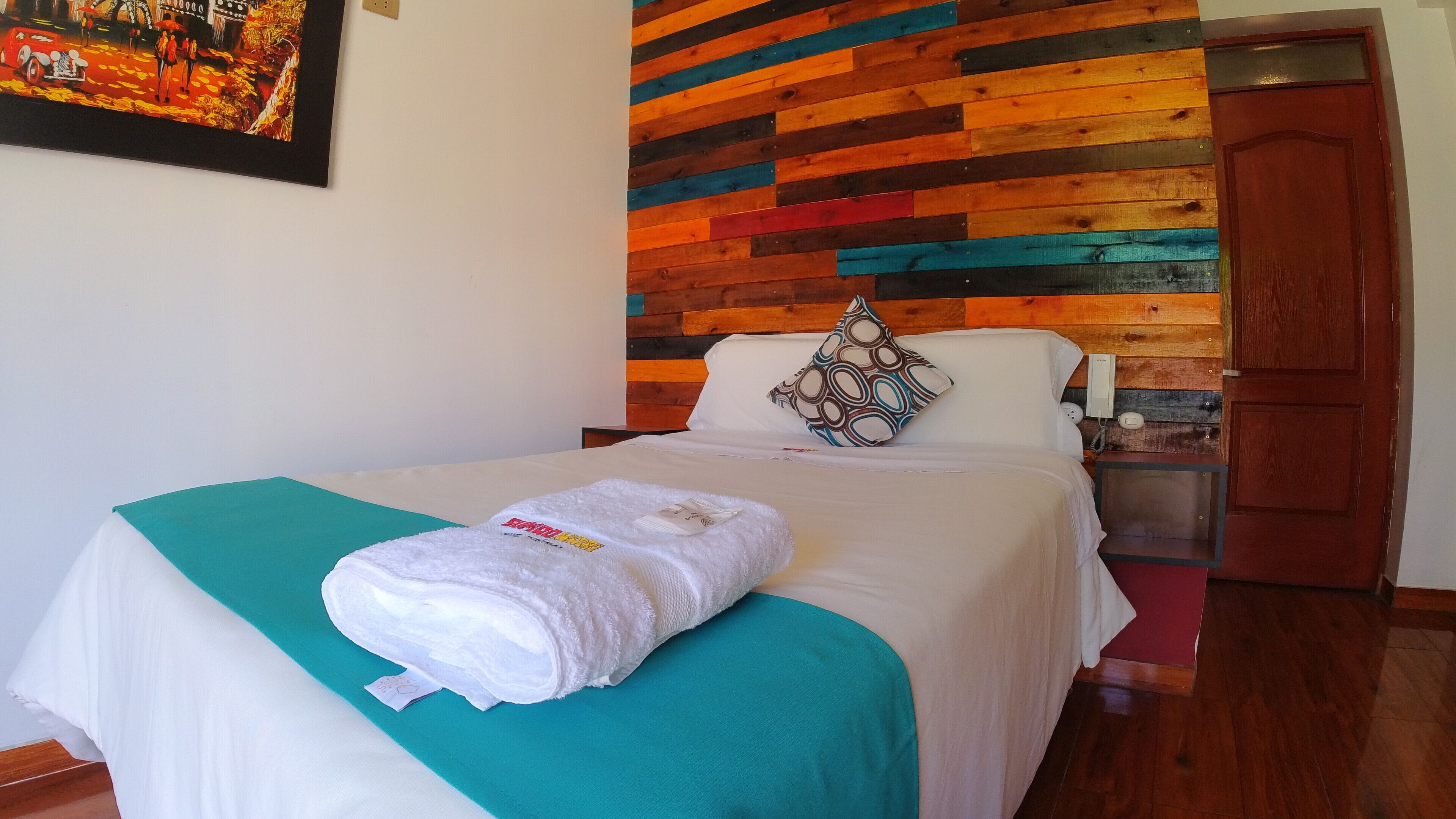 Un lit bien fait, avec une couette blanche et un plaid turquoise, une tête de lit en bois ornée d’un motif coloré, un coussin à motifs et une petite table de chevet sur laquelle se trouve un téléphone.